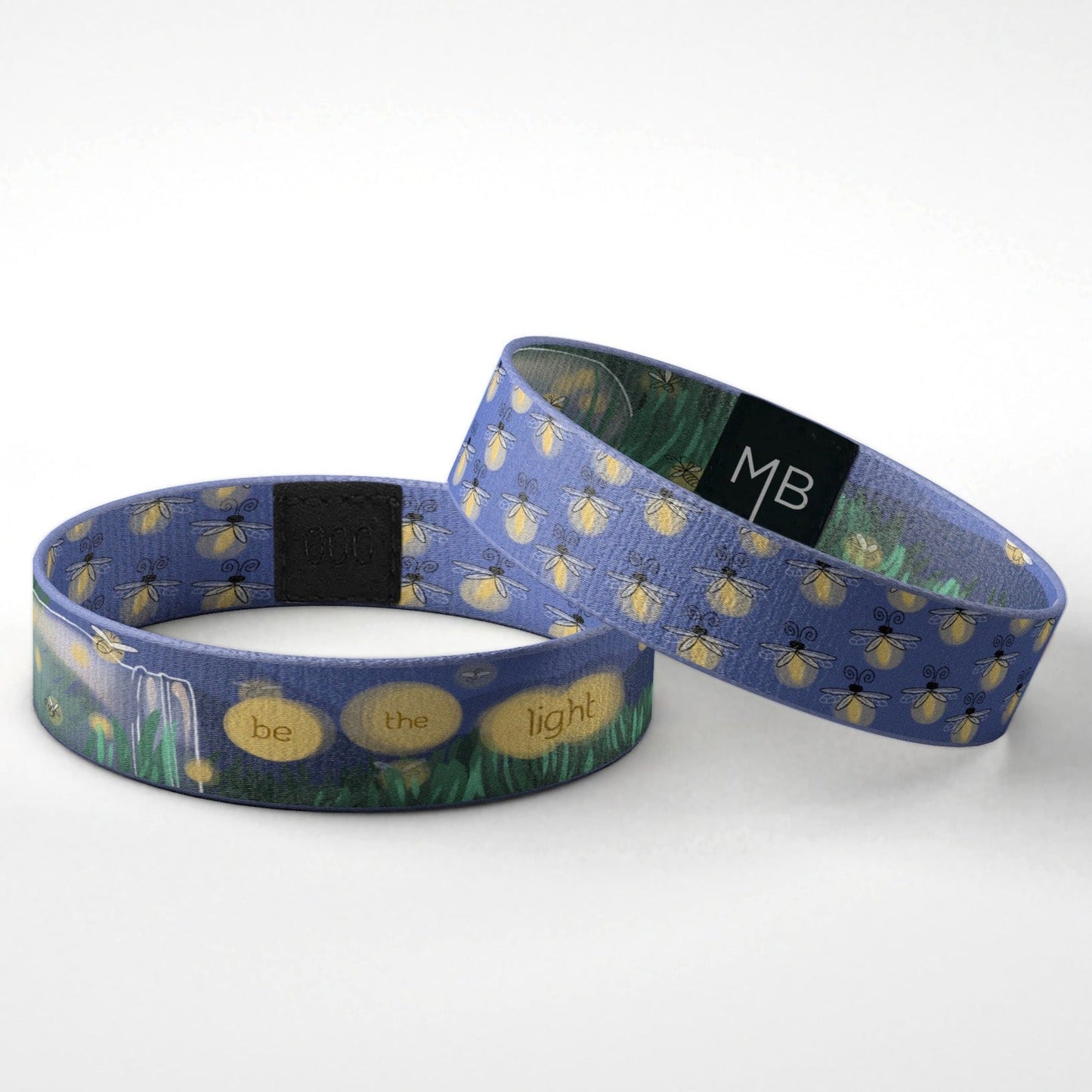 This Moxxie inspirational wristband features little fireflies, or maybe you call them lightning bugs all over in a soft yellow to appear as if they are glowing on a deep cobalt blue background. The text side of this motivational wristband features a Mason jar tipped on its side laying in the grass against a night sky, a grouping of the fireflies illuminates the words “be the light.” The reversible side of this band is cobalt blue and wrapped in a pattern of fireflies. Laying Down View.