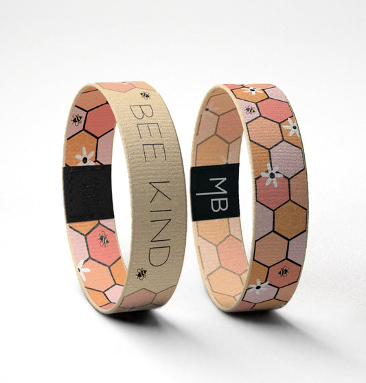 This Moxxie wristband is beautifully lined with pink and peach honeycombs. Both sides of this wristband feature little white daisies sprinkled throughout. The honeycombs & daisies are accompanied by a few mini honey bee’s. The “Bee Kind” side of the band is a warmer, muted version of the reversible side. The muted pale yellow highlights the message of this band. Standing up view.